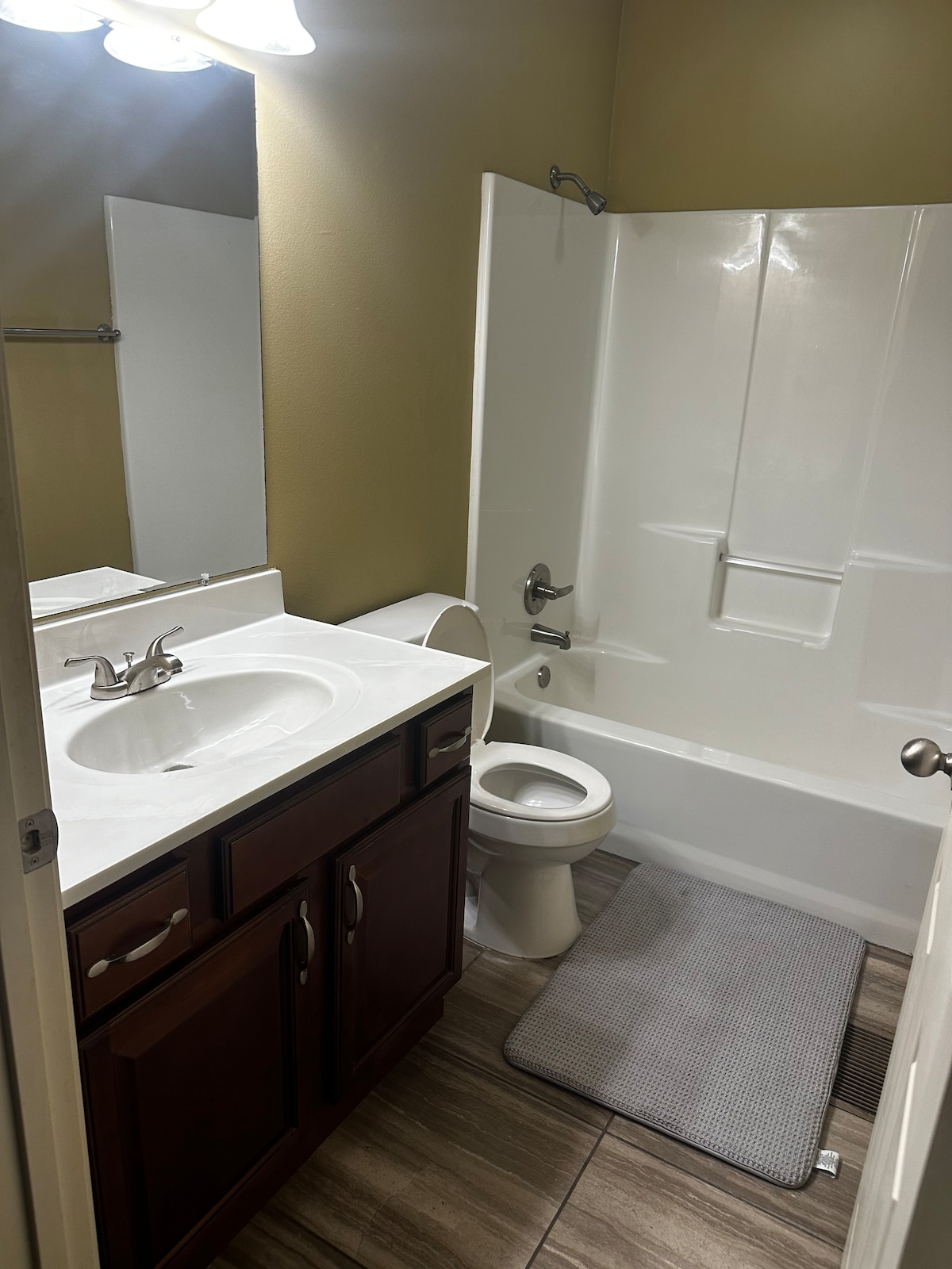 A well-maintained bathroom features a white vanity with a sink, complemented by wooden cabinetry below. A bathtub with a showerhead is present, alongside a toilet and a gray bath mat on tiled flooring. Soft lighting is provided by overhead fixtures.