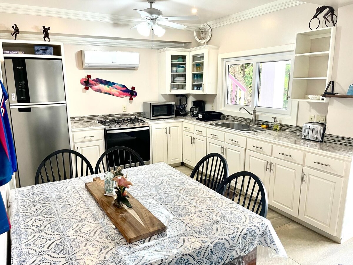 A well-equipped kitchen features modern appliances, including a refrigerator, microwave, and gas stove. A dining table with black chairs sits at the center, accompanied by a decorative centerpiece. Natural light is provided through the window, highlighting the organized cabinets and countertops.