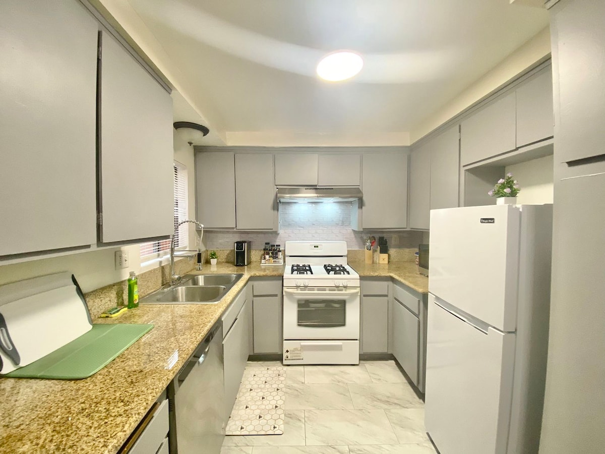 A modern kitchen features sleek gray cabinets and a contrasting white refrigerator. Equipped with a gas stove, sink, and ample counter space, the layout facilitates cooking and meal preparation. Natural light enters through a nearby window, enhancing the functional design.