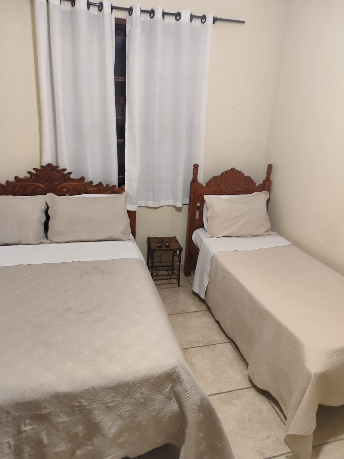 A cozy sleeping area features two wooden beds, one larger and one smaller, adorned with beige bedding. A small nightstand is positioned between the beds, and light filters through a window covered by white curtains, adding a soft ambiance to the room.