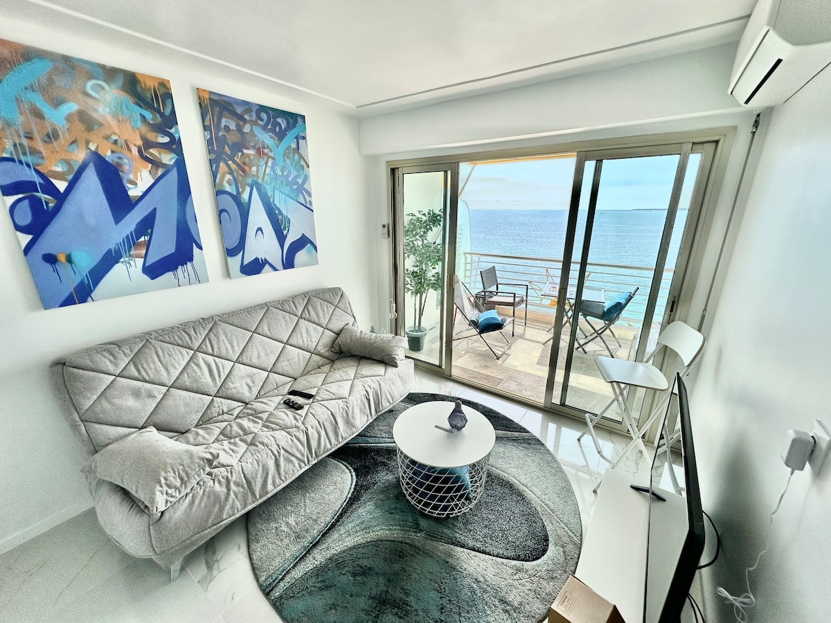 A living area showcases a comfortable, light-colored sofa against a backdrop of vibrant artwork. A glass door opens to a balcony with seating for two, offering views of the Mediterranean Sea. A circular table and a textured rug enhance the space's modern design.