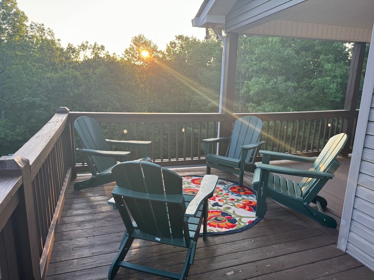 A wooden deck features four green adirondack chairs arranged around a colorful area rug. Morning sunlight filters through the surrounding trees, casting a warm glow over the space, inviting relaxation with views of nature.