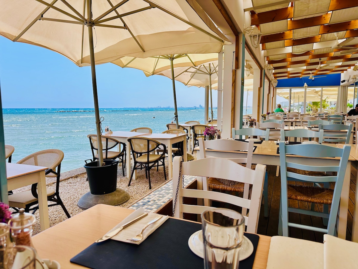A beachfront dining area is displayed, featuring numerous white tables and chairs under large umbrellas. The serene sea is visible in the background, with gentle waves lapping at the shore. The space is bright and inviting, with sunlight enhancing the coastal ambiance.