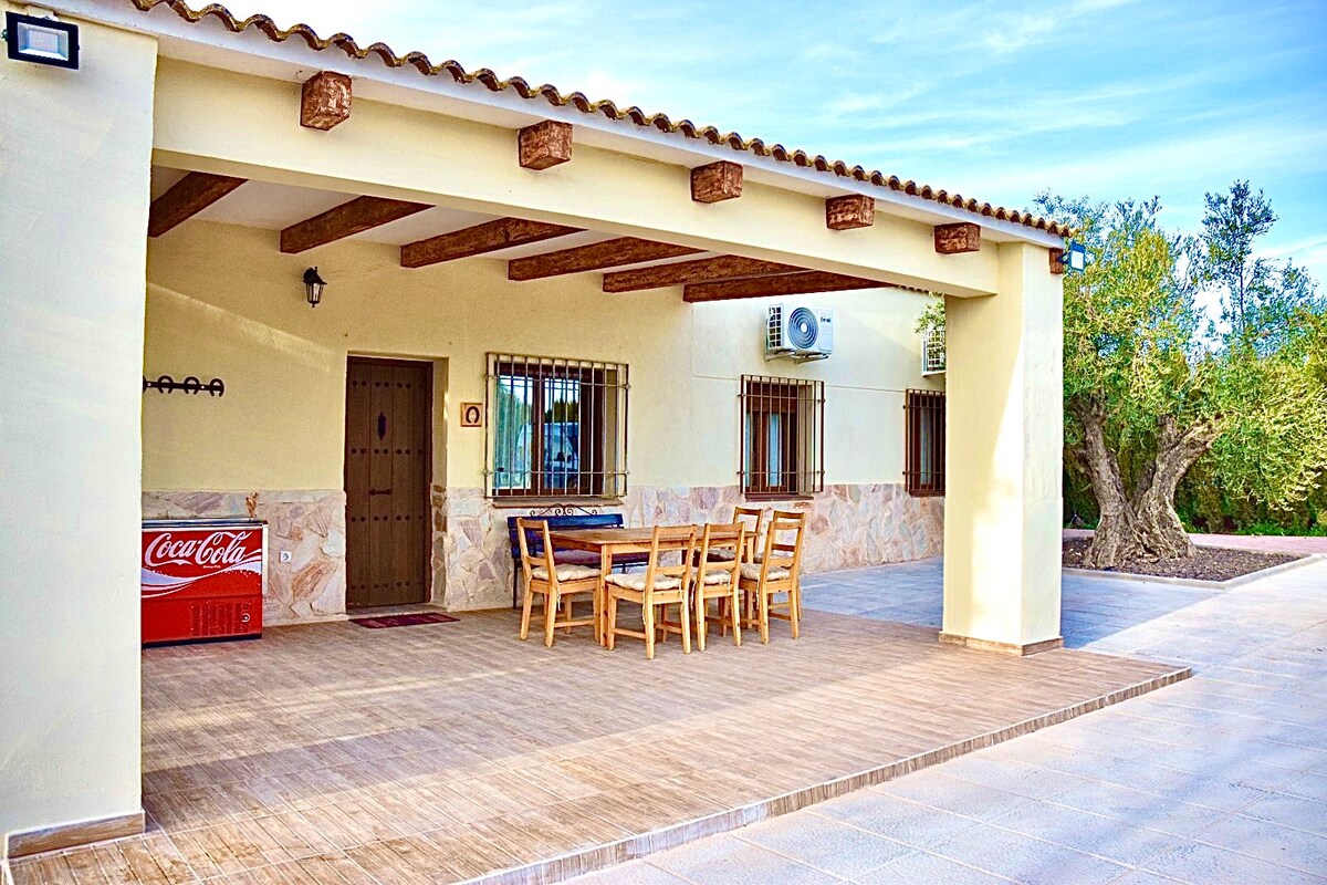 A covered outdoor patio area features a wooden dining table surrounded by several chairs. The floor is tiled, and a wall-mounted air conditioning unit is visible. A classic Coca-Cola cooler is positioned against the wall, with olive trees visible in the background.