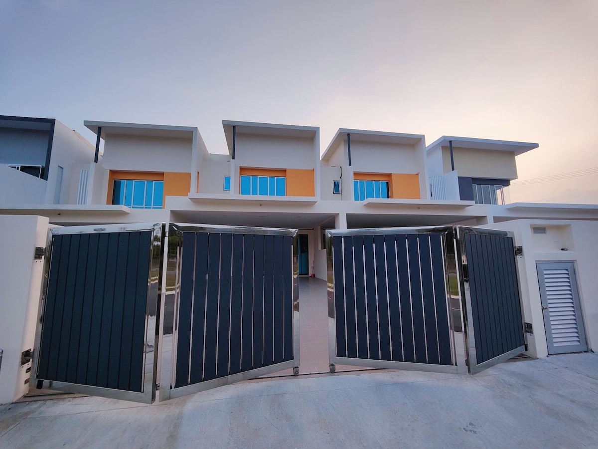 The exterior view of a modern building features a pair of metallic gates opening toward a spacious entrance. The building showcases a minimalistic design with large windows, orange accents, and a neutral color palette, set against a soft evening sky.
