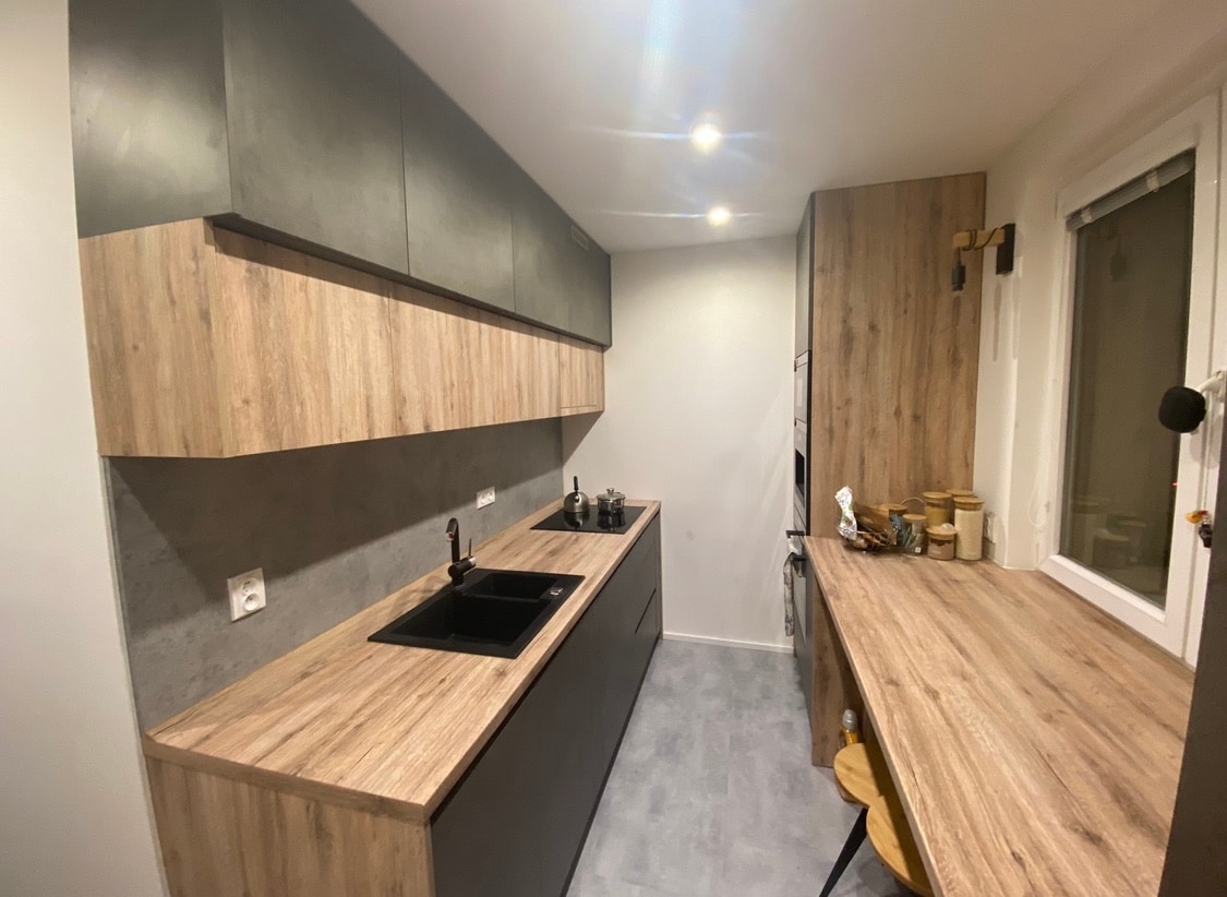 A modern kitchen is presented with sleek cabinetry in a combination of wood and dark finishes. A black sink is integrated into the countertop, complemented by a workspace featuring a wooden surface and stool. Soft lighting enhances the streamlined design.