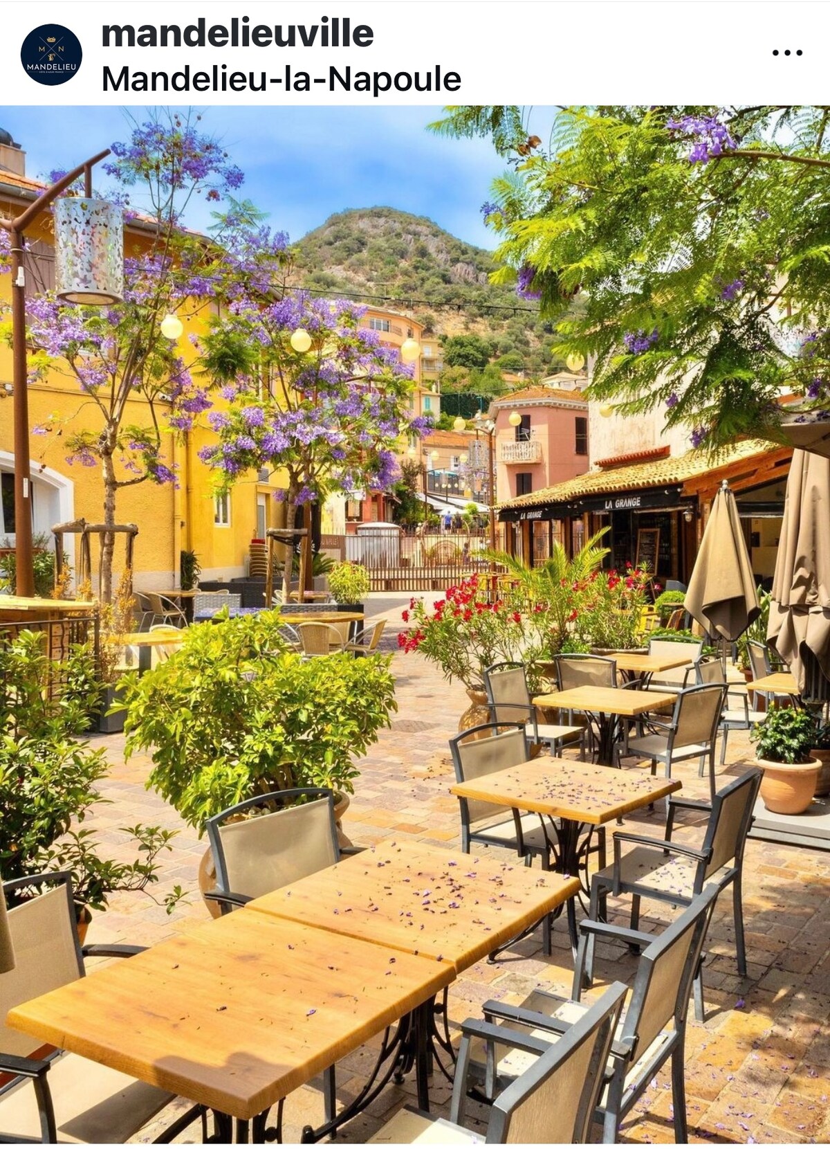 A picturesque street is lined with blooming purple flowers and colorful buildings. Several outdoor tables and chairs are arranged under the sun, inviting guests to enjoy the vibrant atmosphere of the area. A hillside is visible in the background, enhancing the lively scene.