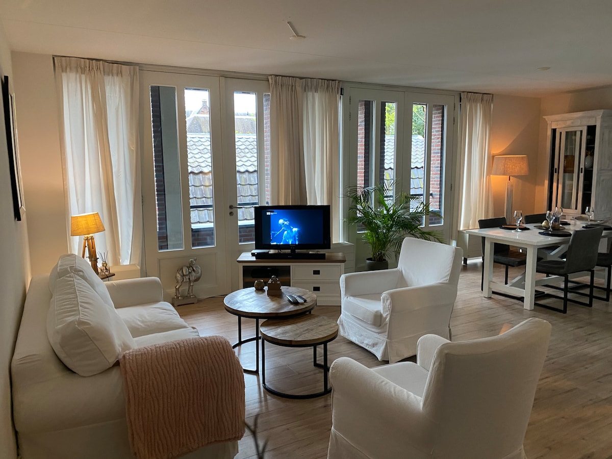 A spacious living area is illuminated by natural light from large windows. Soft, neutral furnishings include a white sofa and armchairs, with a round wooden coffee table at the center. A television and a plant are positioned nearby, and a dining table can be seen in the background.