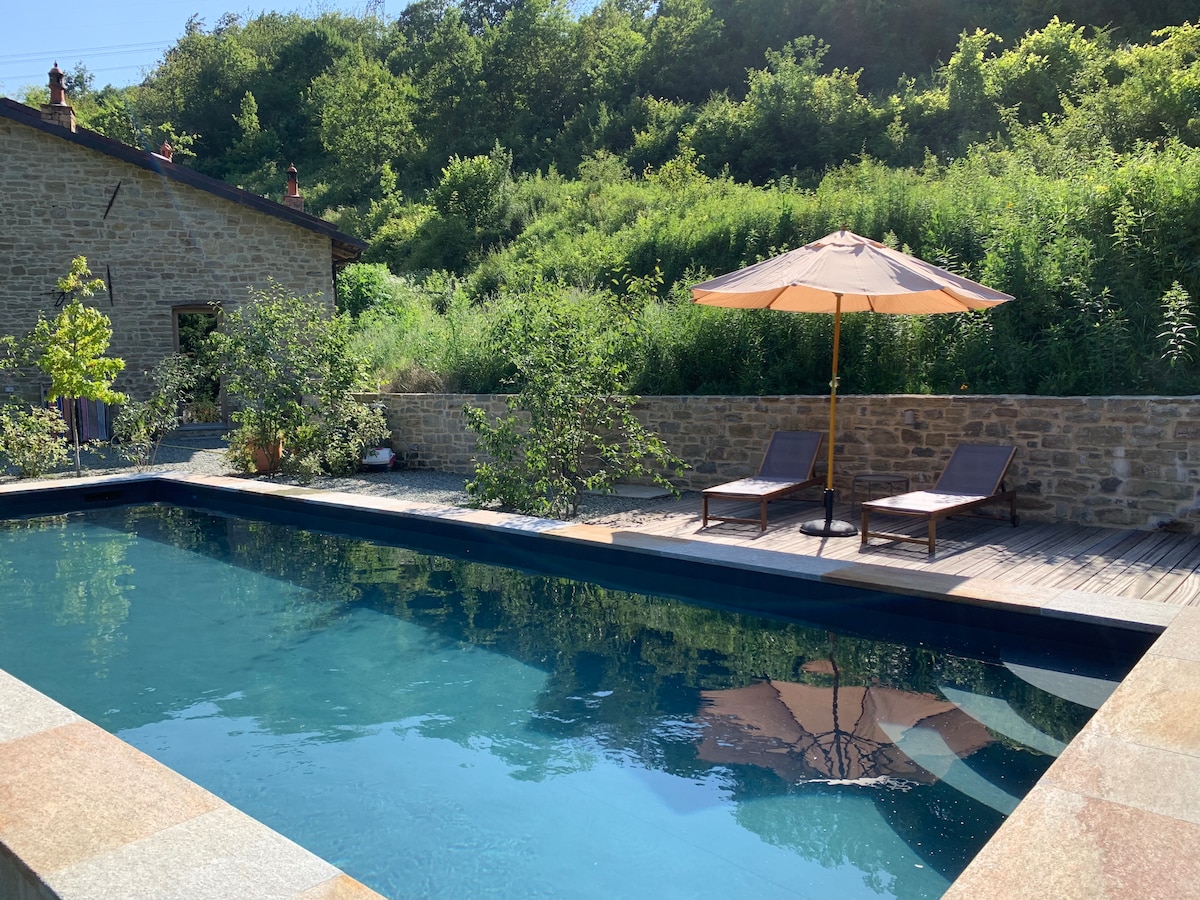 A private swimming pool is framed by stone walls and lush greenery. Two lounge chairs sit on a wooden deck beside the water, complemented by a large umbrella for shade. Sunlight reflects off the serene surface of the pool, enhancing the peaceful setting.