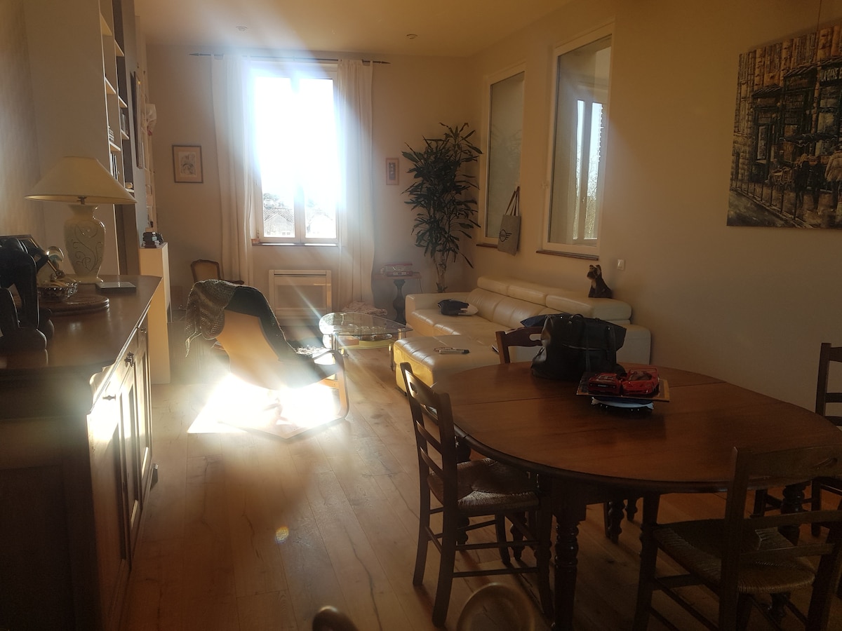 A bright living space features a wooden dining table surrounded by several chairs. A white leather couch is positioned near a large window, with sunlight illuminating the area. A decorative plant adds a touch of greenery, and art is displayed on the walls.