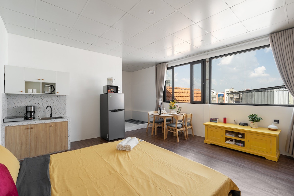 A spacious living area features a cozy bed covered in a yellow blanket. A kitchenette with modern appliances is visible in the corner, while a dining table for four is positioned near large windows that allow natural light to fill the space.