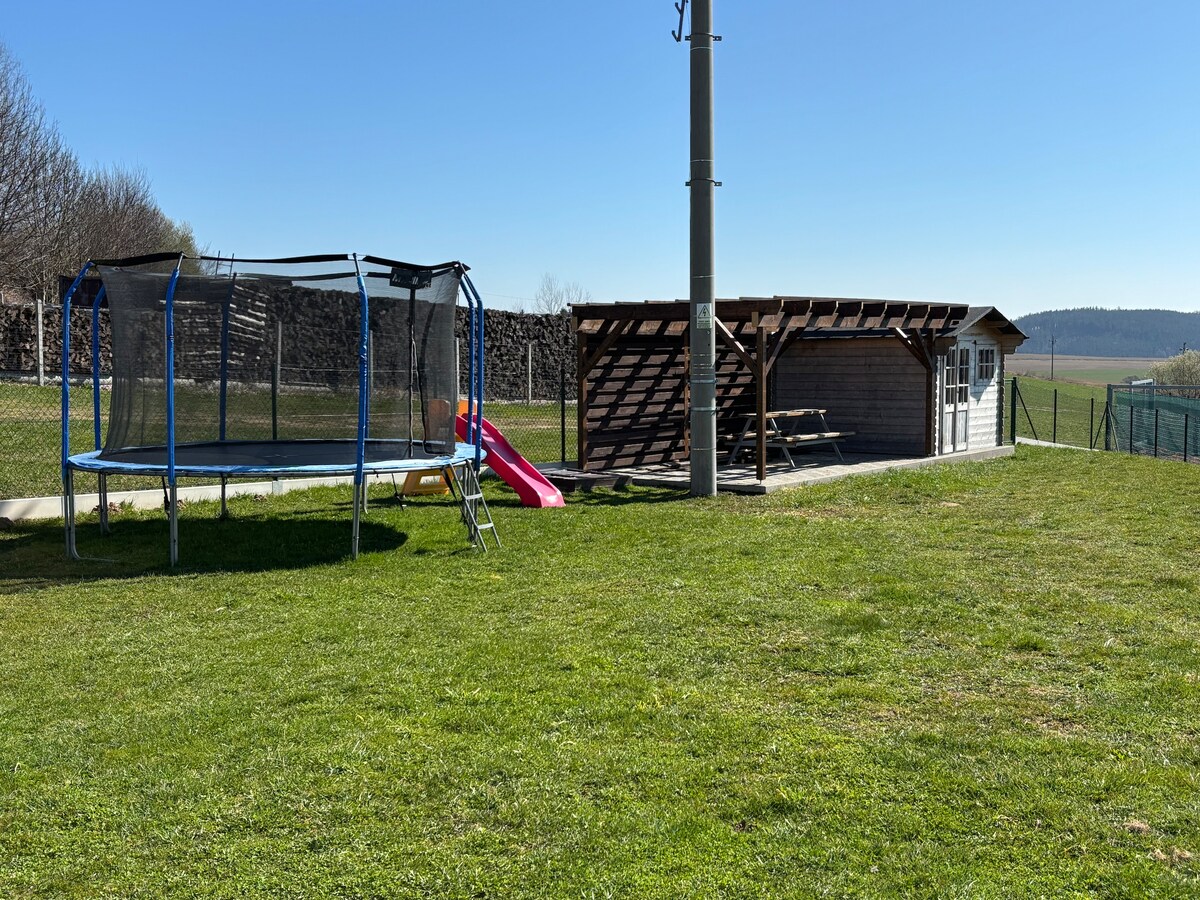 An outdoor area features a grassy lawn with a trampoline and a small slide nearby. A wooden shelter offers seating and is positioned alongside a rustic building. Clear skies and a distant landscape can be seen in the background.