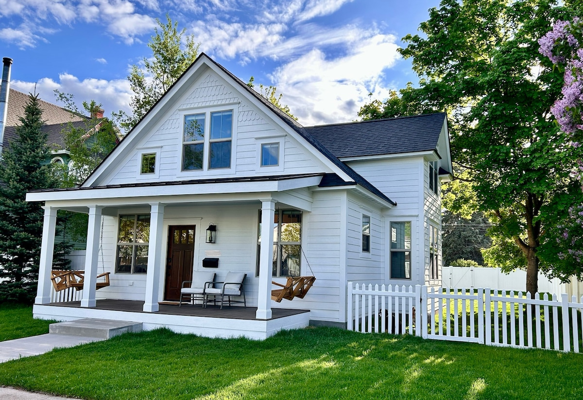 The charming exterior reveals a classic white wooden facade with a covered porch adorned with two swinging chairs. Lush green grass surrounds the home, complemented by a white picket fence. Tall trees provide shade, and a clear blue sky enhances the inviting atmosphere.
