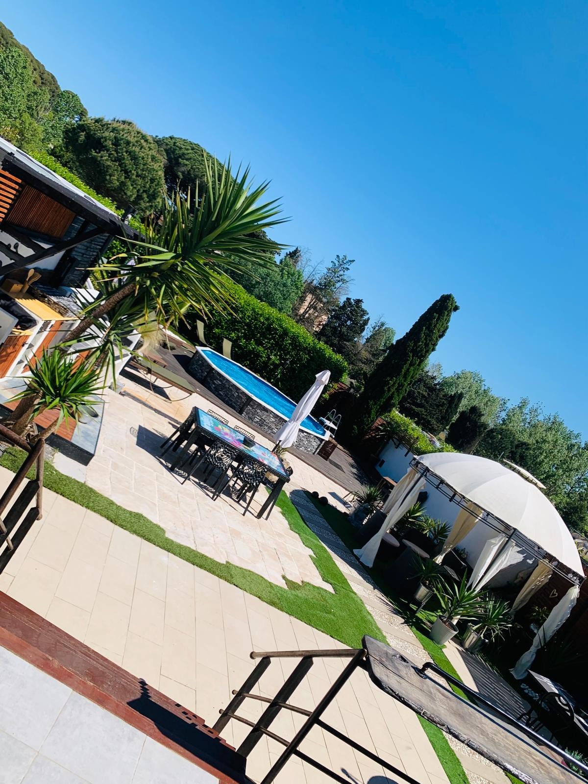 An outdoor area showcases a spacious patio with a dining table and comfortable seating arrangements. The clear blue sky complements a shimmering pool and a nearby gazebo, surrounded by greenery. Sun loungers are placed for relaxation, enhancing the inviting ambiance of the space.