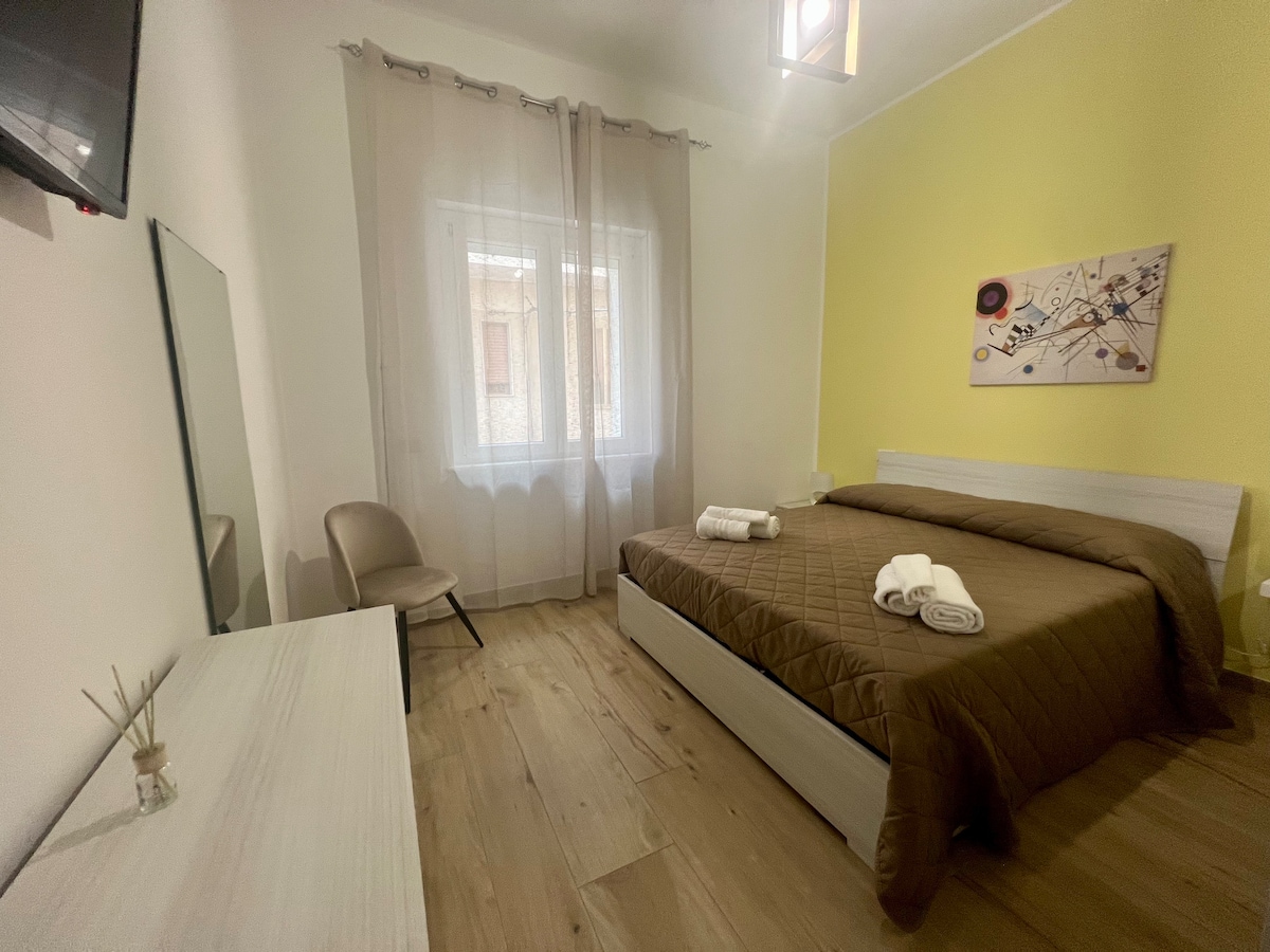 A bright sleeping area features a spacious bed with a brown quilt, complemented by white linens. A decorative wall piece adds color to the yellow accent wall. Natural light enters through a window adorned with sheer curtains, and modern furnishings include a mirror and a chair.