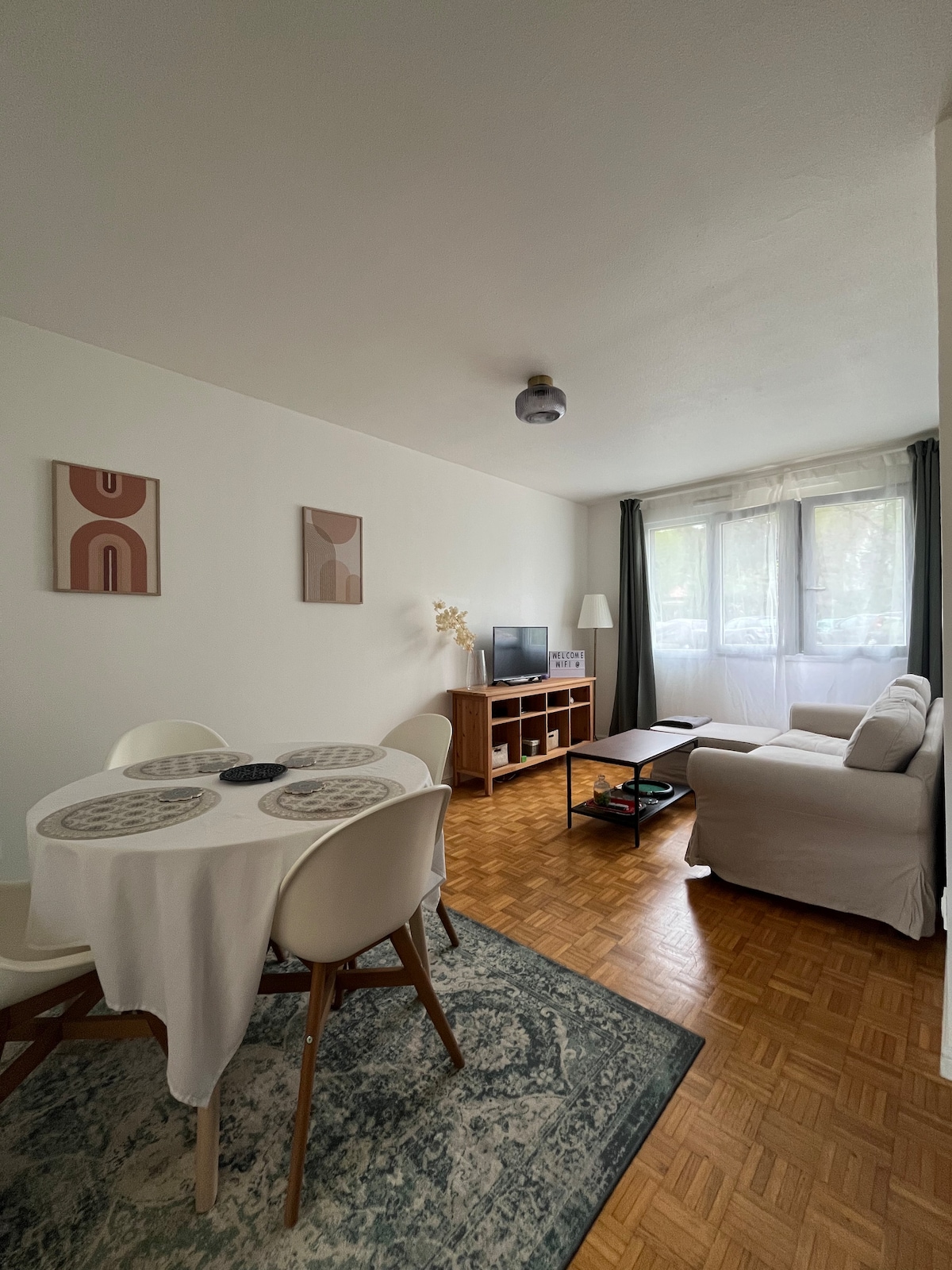 A modern living area is depicted, featuring a light-colored sofa and a coffee table on a textured rug. A dining table with white chairs sits nearby. Natural light filters through large windows, illuminating the space and enhancing the minimalist decor on the walls.