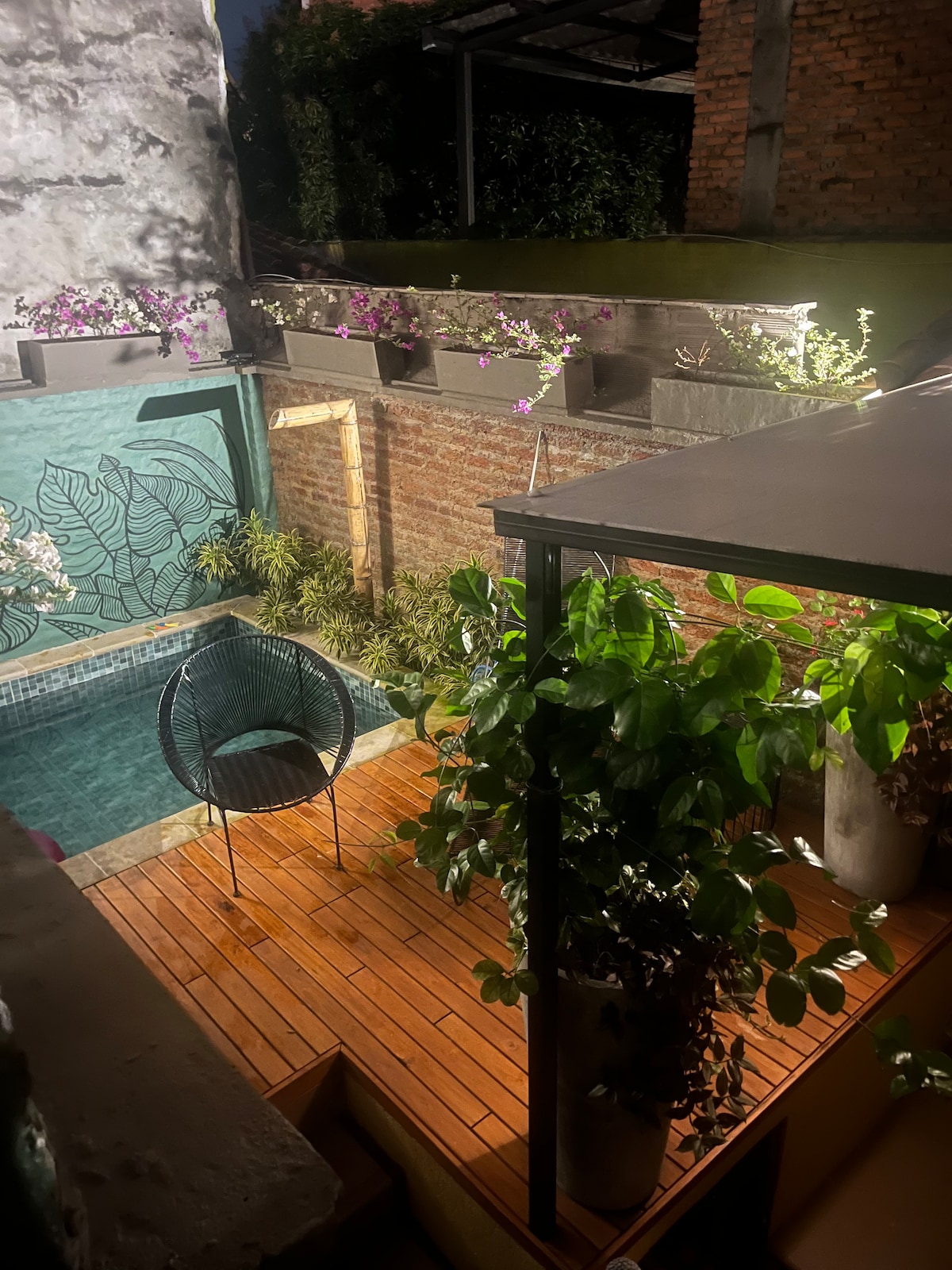 A small pool is surrounded by a wooden deck, featuring an outdoor chair and potted plants. A mural with botanical elements is displayed on a nearby wall, while flowers bloom in planters above, contributing to a serene outdoor environment.