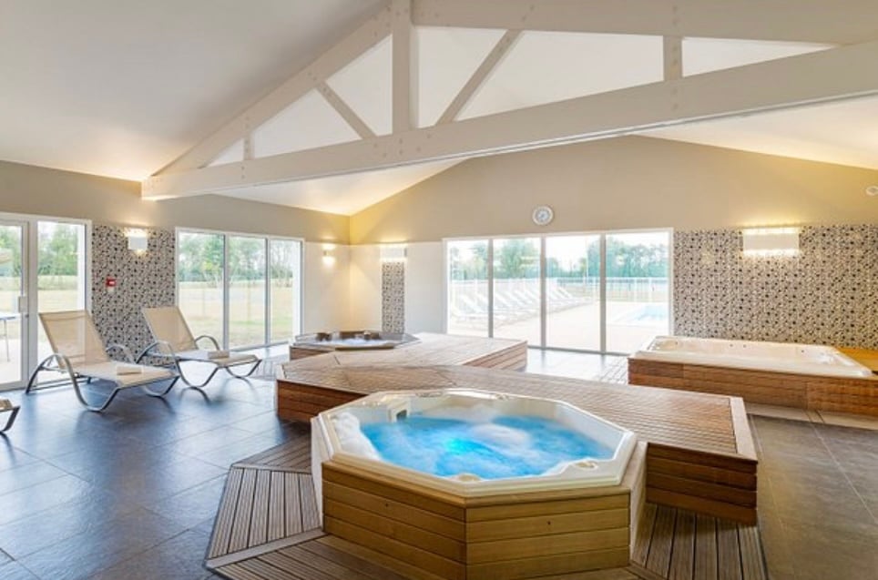 The spa area is designed with a modern aesthetic, featuring multiple hot tubs surrounded by wooden decking. Large glass windows allow natural light to fill the space, with views of the outdoor pool area visible beyond. Lounge chairs are arranged for relaxation.