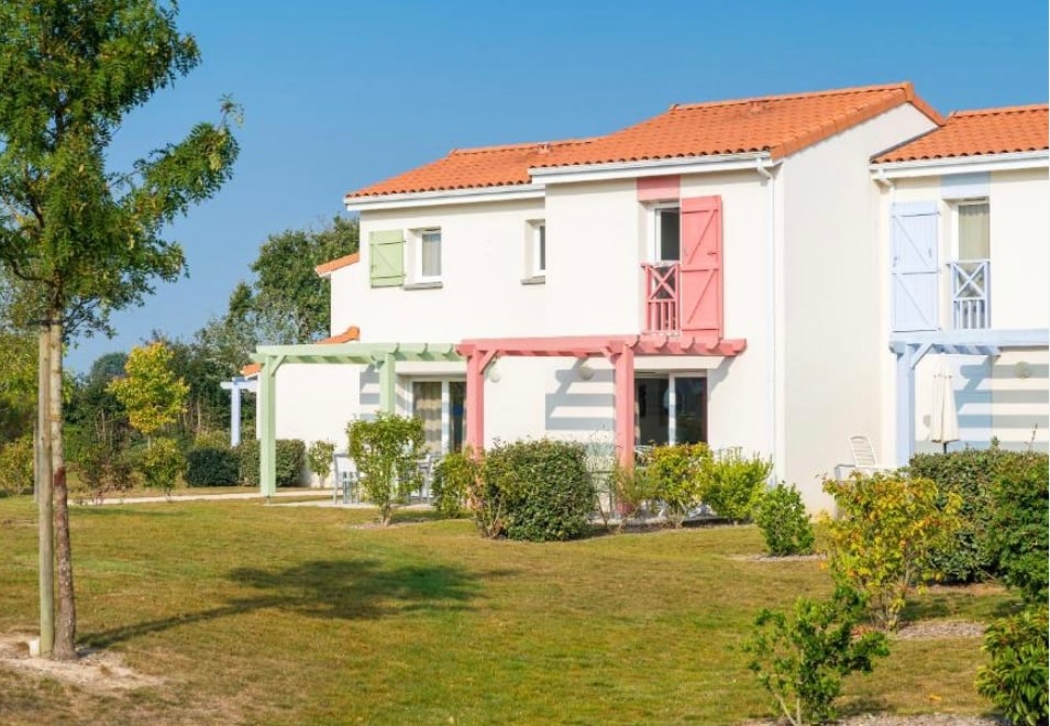 A charming two-story residence features a bright exterior with colorful shutters in pink and green. Lush greenery surrounds the building, complemented by well-maintained shrubs and trees. The blue sky above enhances the cheerful atmosphere of this peaceful country setting.