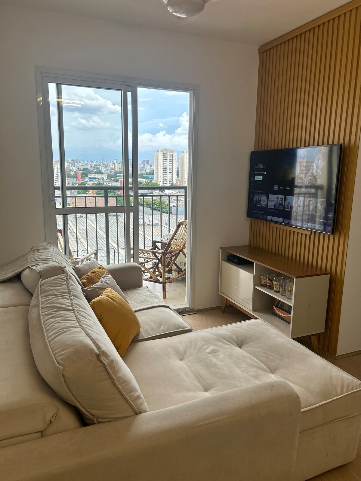 A cozy seating area features a light-colored sofa complemented by yellow accent pillows. A large window provides a view of the cityscape, allowing natural light to fill the space. A wall-mounted television is positioned near a modern media console, creating a comfortable relaxation spot.