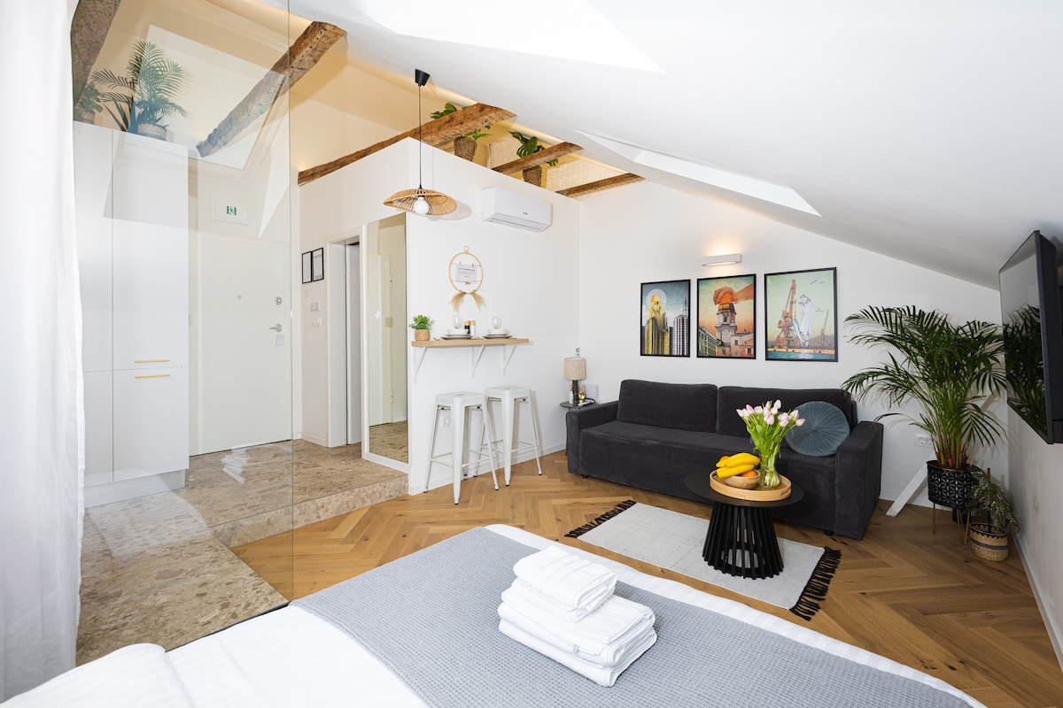 A bright studio apartment features a spacious bed covered with fresh linens. A cozy seating area includes a dark sofa beside a round table with fruit. Neatly arranged artwork adorns the walls, while natural light floods through skylights and a window.