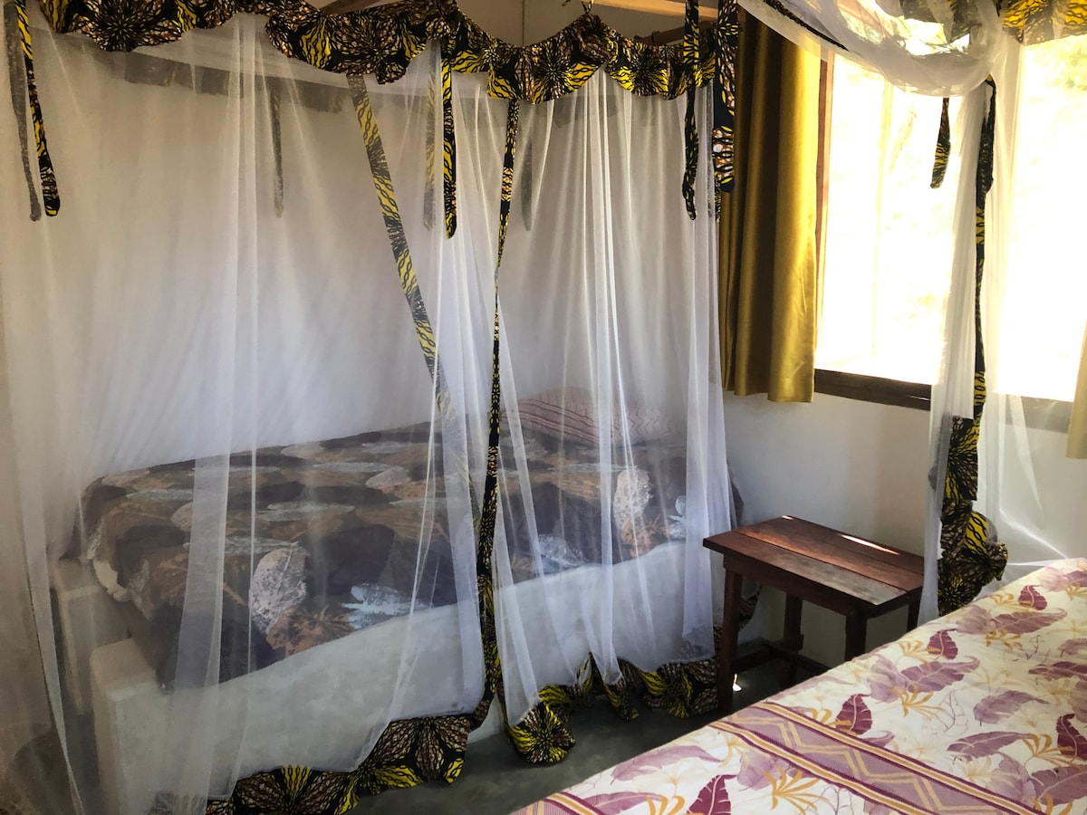The room features two large single mattresses, each with a mosquito net draped above. A small side table is positioned between the beds. Natural light enters through a window with light curtains, creating a bright and airy space.