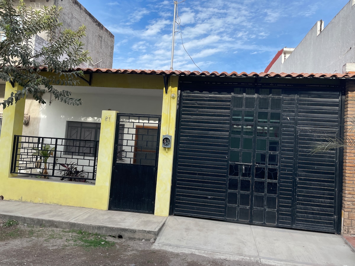 The exterior of a yellow house features a gated entrance with a black metal gate and a contrasting black garage door. A well-maintained pathway leads to the front door, framed by a small garden. Clear blue skies provide a bright backdrop.