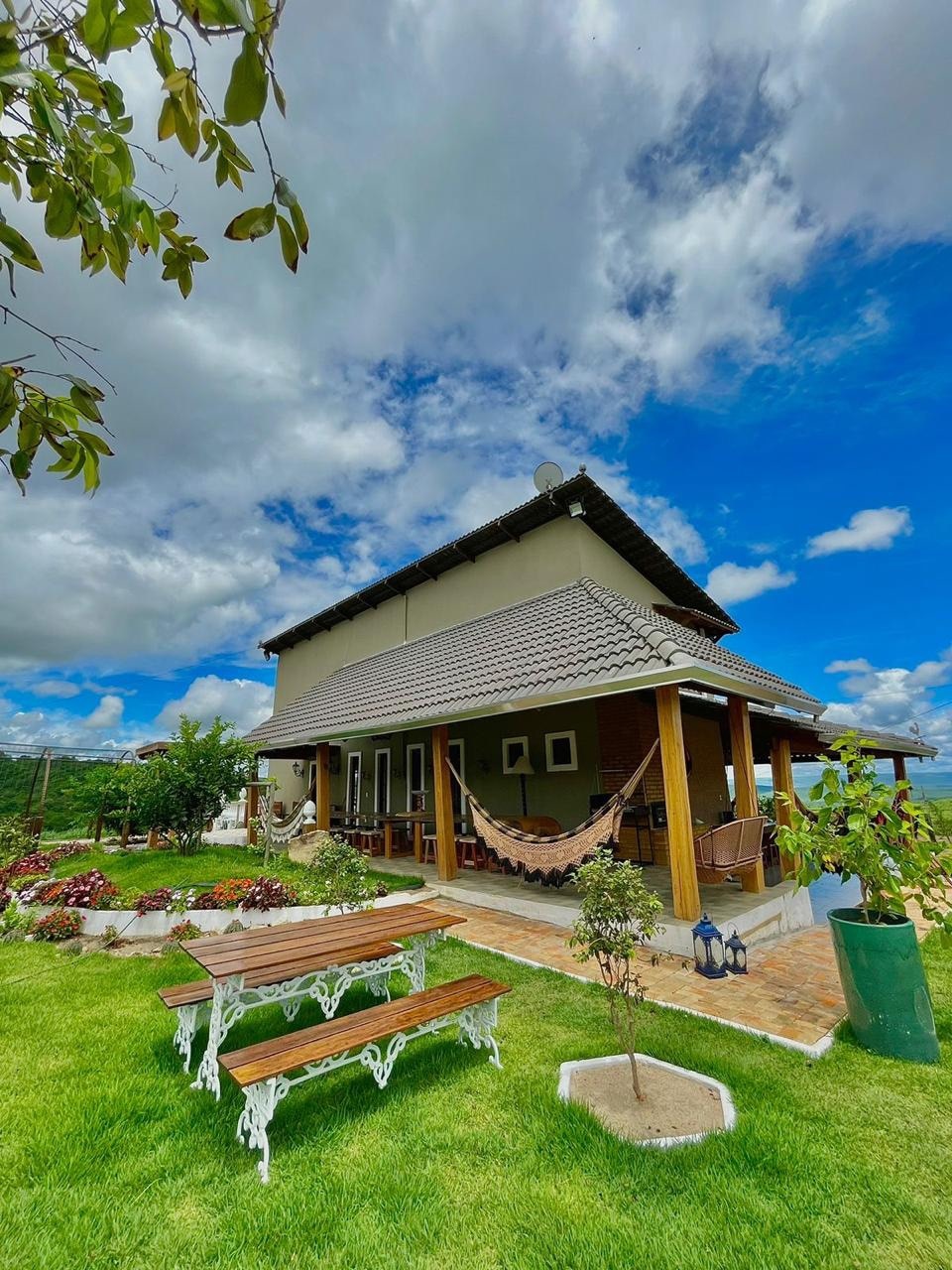 A charming two-story house is surrounded by lush greenery and colorful flower beds. A spacious porch with a hammock invites relaxation, while a wooden picnic table provides outdoor dining space. The clear blue sky complements the architectural features of the home.