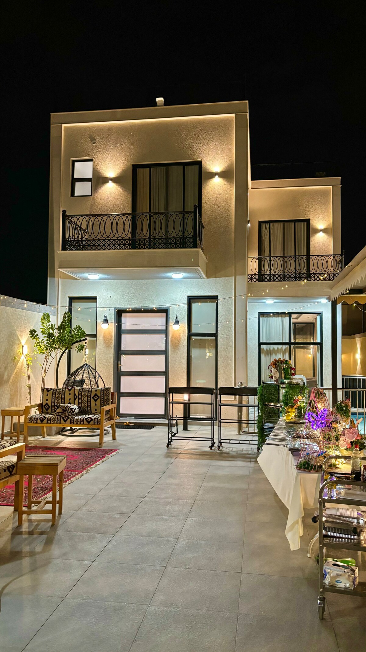 A modern two-story building is illuminated at night, showcasing a bright entrance with large glass doors and balcony features. Outdoor seating arrangements with wooden chairs and decorative elements are visible, enhancing the inviting atmosphere of the patio area.