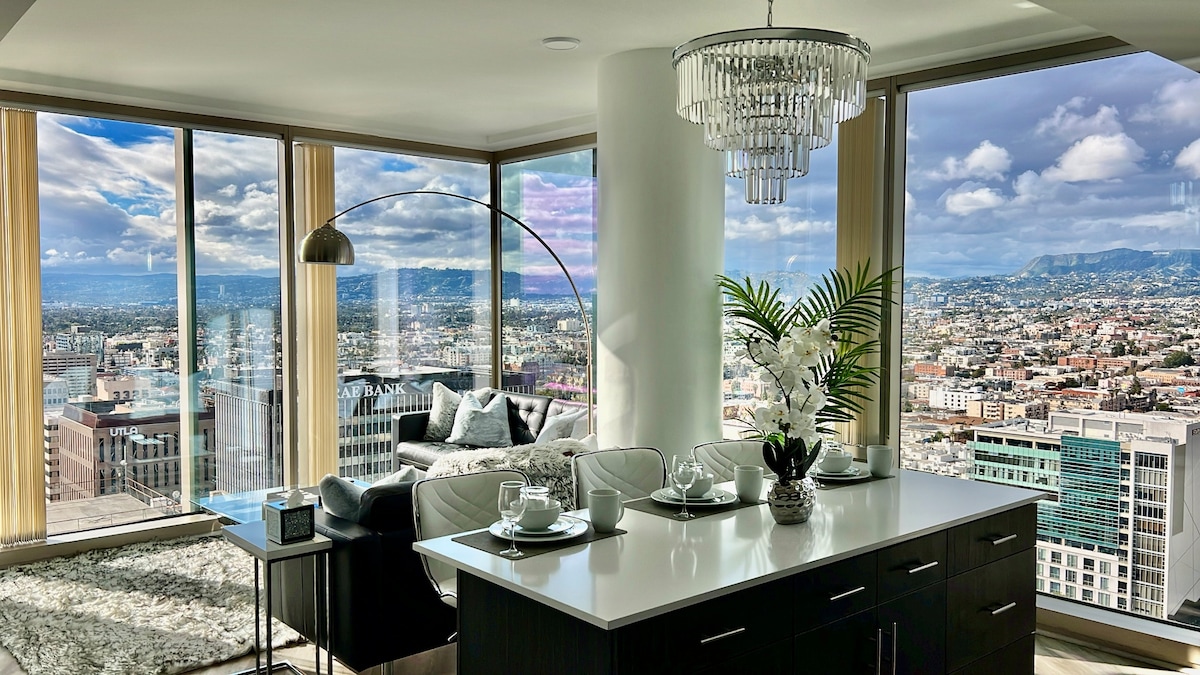 A modern living area is showcased, featuring floor-to-ceiling windows that provide expansive views of Los Angeles. A sleek white countertop is set for dining, adorned with plates and decorative accents. Plush seating and a decorative plant add to the contemporary ambiance.