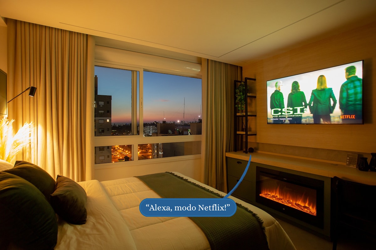 A well-appointed sleeping area features a spacious bed with green and white linens. An electric fireplace adds warmth, while a wall-mounted SmartTV displays a show. Large windows reveal a colorful sunset over the city skyline.
