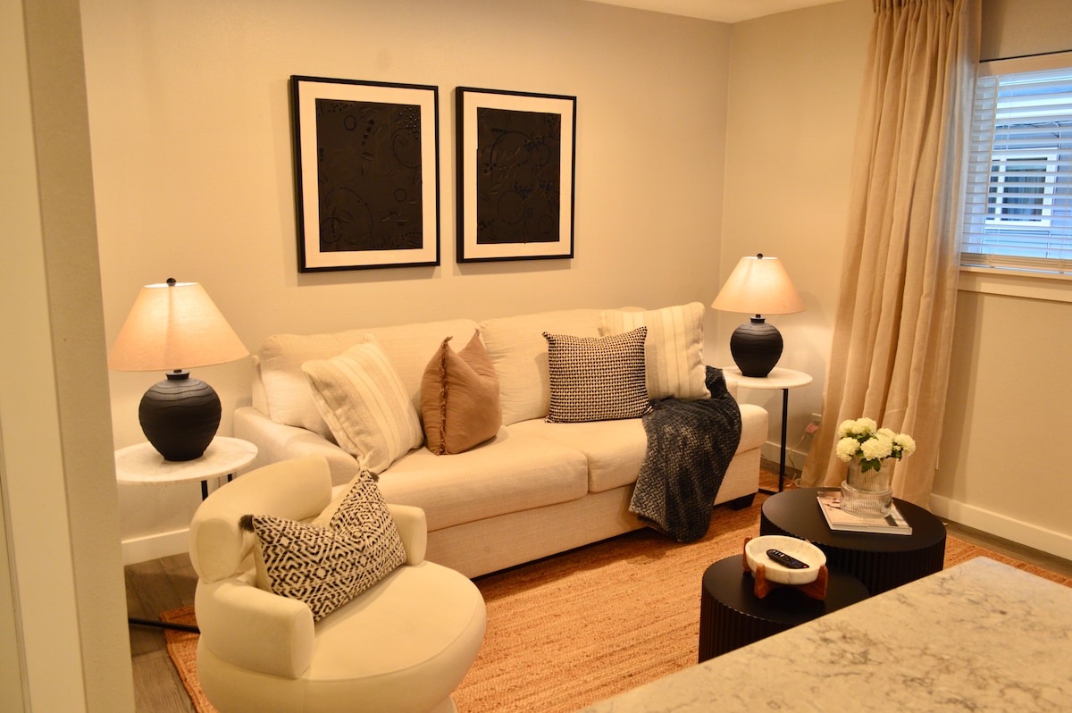 A bright living area features a light-colored sofa adorned with decorative pillows, complemented by two stylish side tables. Soft lighting is provided by table lamps on either side, while a textured rug adds warmth to the space. Two framed artworks enhance the modern aesthetic.