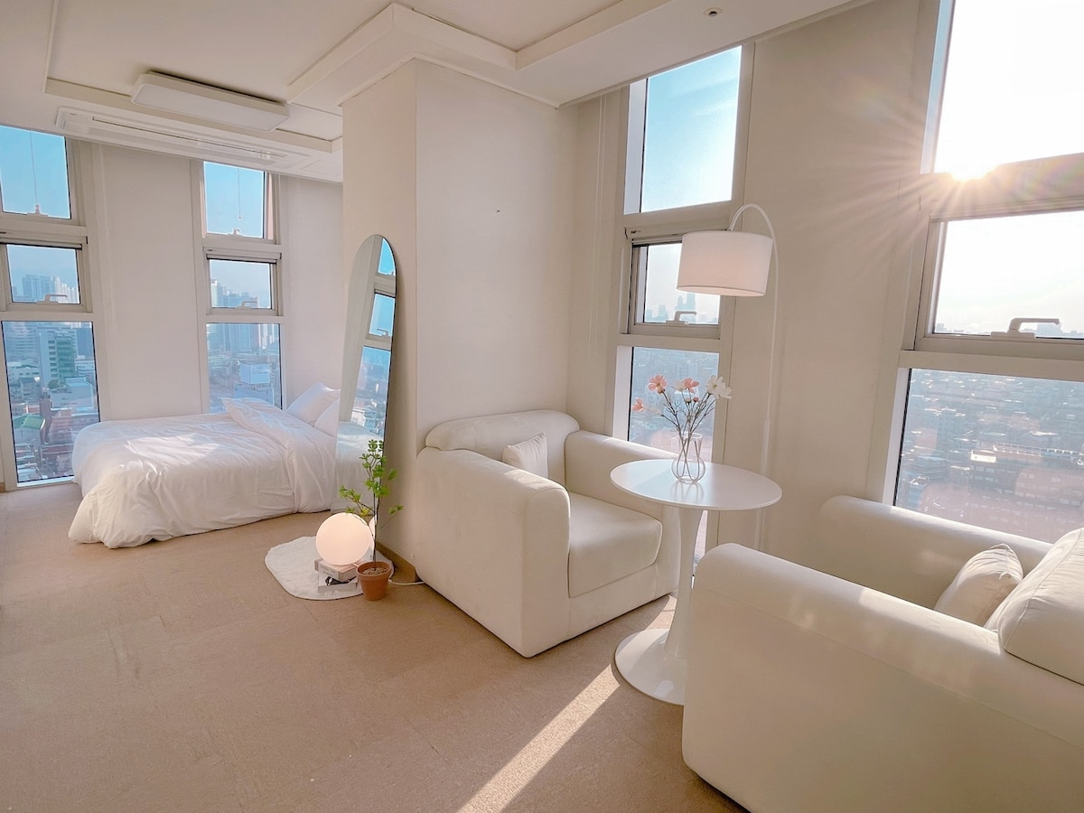 A well-lit open space features large windows that allow natural light to fill the room. A cozy sleeping area contains a spacious bed, while a seating arrangement with two white chairs and a small table invites relaxation. Subtle decor elements add to the serene ambiance.