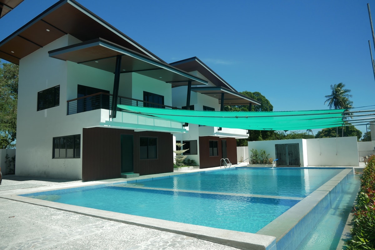 A spacious shared swimming pool is observed in the foreground, accompanied by two modern residential buildings featuring large windows and balconies above. A green canopy provides shade over part of the pool area. Lush greenery is visible in the background, complementing the serene setting.