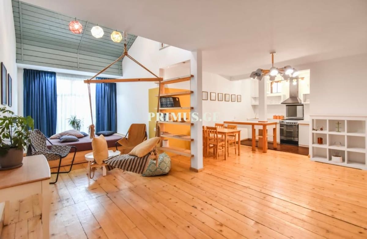 A spacious living area is illuminated by natural light through large windows. A hanging chair and a cozy seating arrangement are visible, alongside a dining table and modern kitchen appliances. The warm wooden floor enhances the room's inviting vibe.