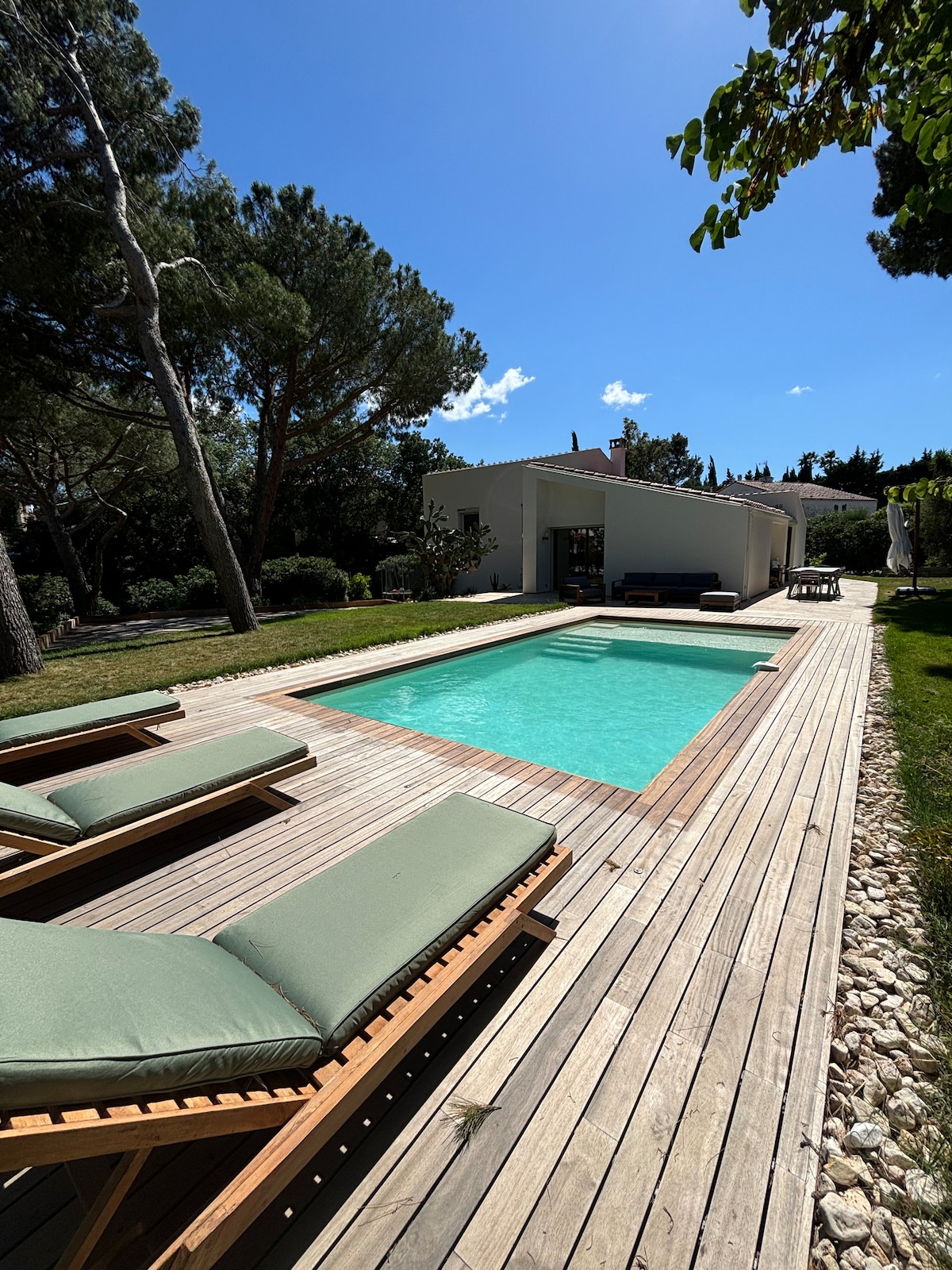 A serene outdoor space features a rectangular pool surrounded by a wooden deck. Green lounge chairs invite relaxation under a clear blue sky. Lush greenery and trees provide a natural backdrop, enhancing the tranquil atmosphere of the setting.