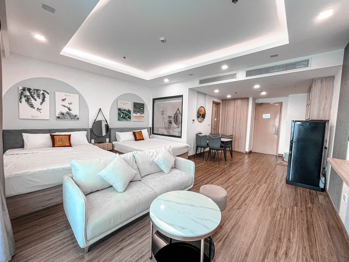 The spacious room is furnished with a large, comfortable sofa and a round marble table. Two beds are positioned against the wall, accompanied by framed artwork. A dining area with a table and chairs is visible, and a kitchen area is located to the right.