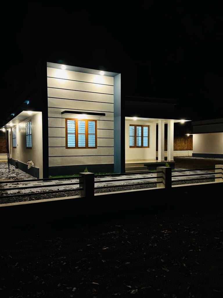 The exterior of a villa is illuminated at night, featuring a modern design with horizontal siding. Bright white lights highlight the structure, and multiple windows allow glimpses inside. A path leads toward the entrance, complemented by a neat green border.