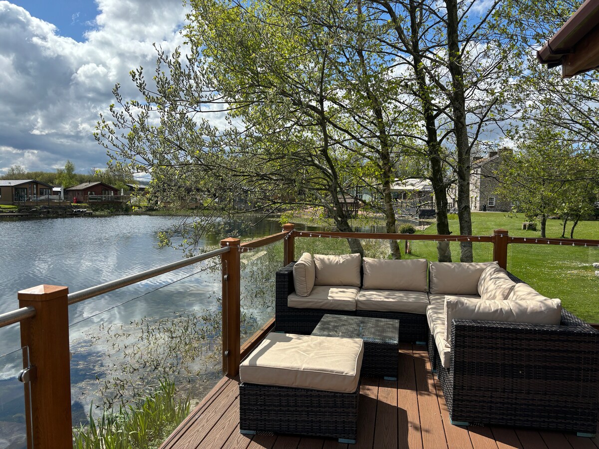 An outdoor seating area features a dark wicker sofa with soft cushions arranged around a low coffee table. The deck overlooks a serene lake, with vibrant green trees framing the view and grass in the background, inviting relaxation.