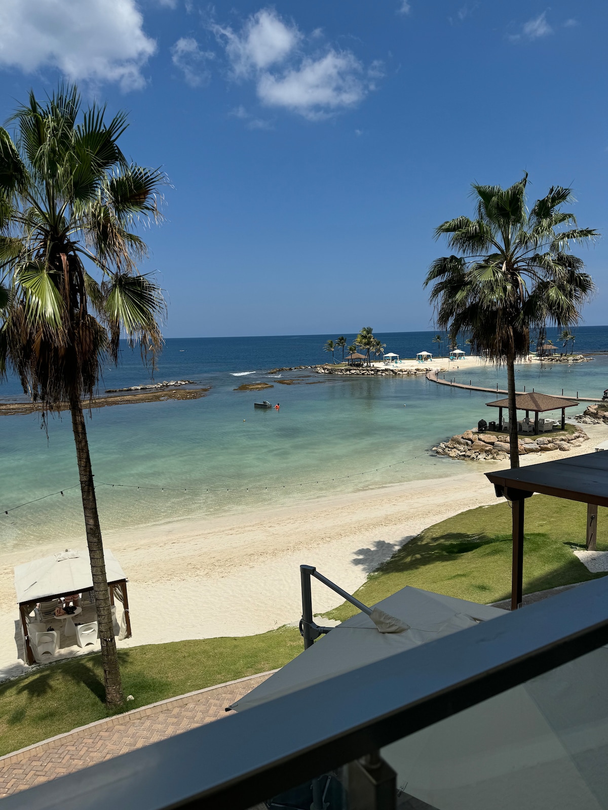 A serene beach view is presented, with gentle waves lapping against the shore. Palm trees sway under a clear blue sky, framing a calm lagoon. A sandy beach leads to a rocky area, where small boats are moored, and shaded pergolas provide relaxation spots.