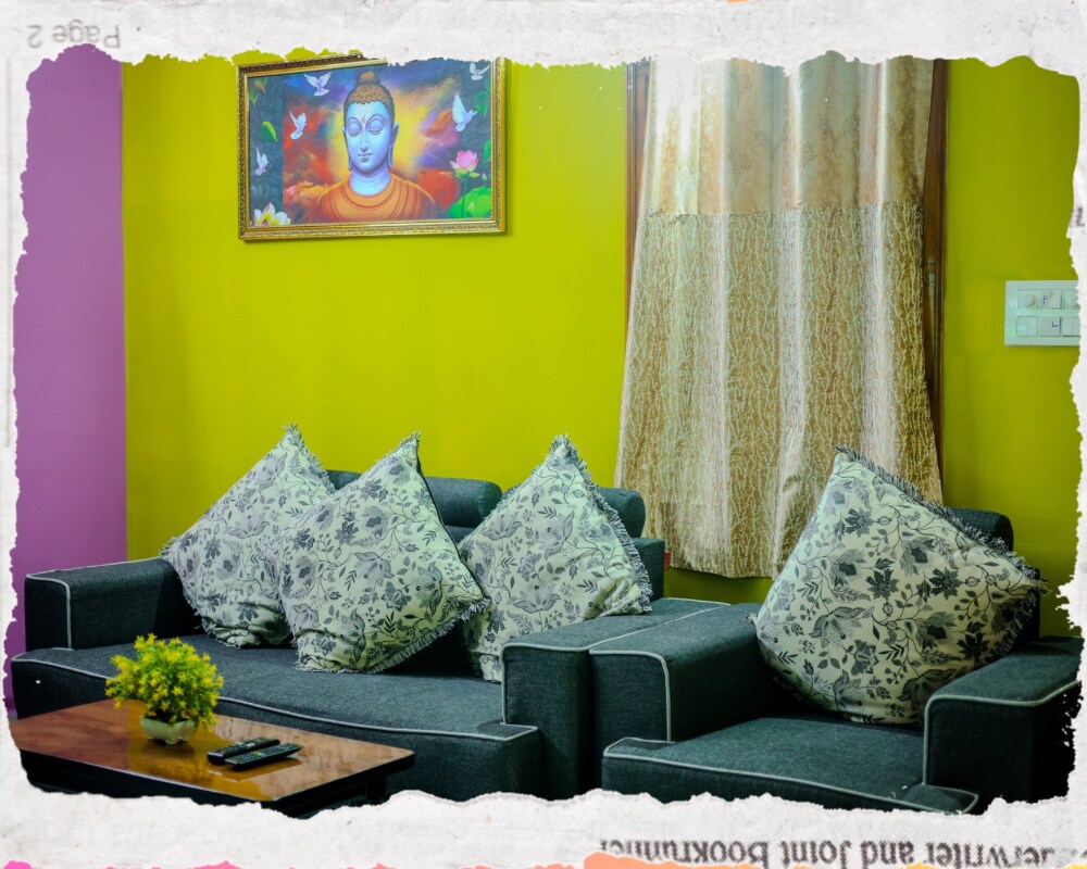 A comfortable living area is presented with a dark upholstered sofa adorned with decorative pillows. A wooden coffee table sits in front, while a framed art piece hangs on a vibrant green wall. Natural light filters through a window draped with soft curtains.