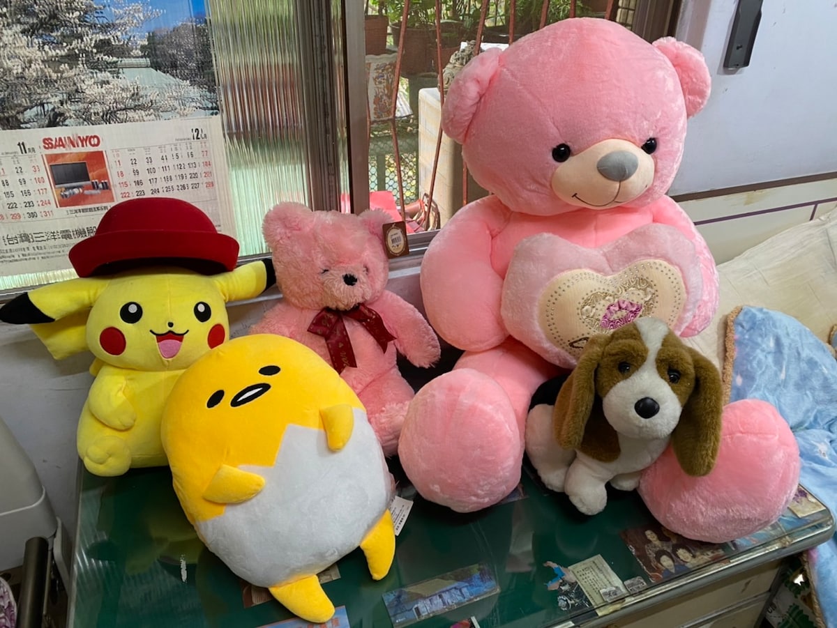 A collection of plush toys is displayed on a glass table. A large pink bear with a heart on its chest sits alongside a smaller pink bear wearing a bow. Other characters include a yellow figure with a skeptical expression, a dog plush, and a cheerful Pikachu, creating a playful atmosphere.