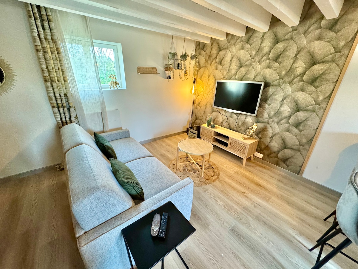 A spacious living area features a light-colored sofa adorned with pillows, positioned across from a wall-mounted television. Natural light filters through a nearby window, complemented by decorative plants and textured wallpaper, creating a calm and welcoming environment.