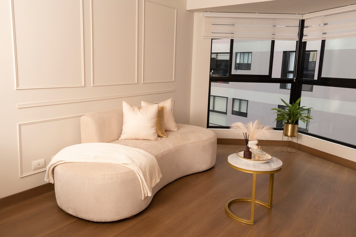 A curved, plush sofa in a neutral tone is positioned near large windows, allowing natural light to fill the room. A round side table with a marble top and golden base complements the space, while decorative elements like soft pillows and a potted plant add subtle charm.