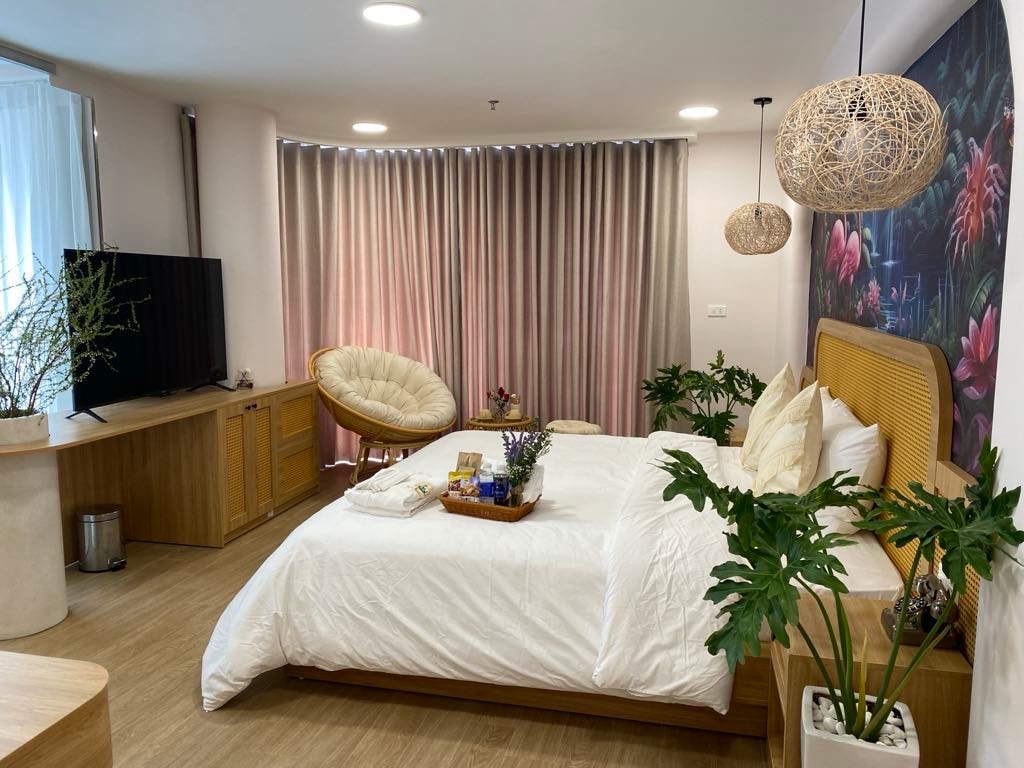 A bright and airy bedroom features a spacious bed dressed in white linens, complemented by decorative pillows. A round, cushioned chair is positioned beside a wooden TV unit. Soft lighting is provided by hanging pendant lamps, while plants add a touch of greenery.