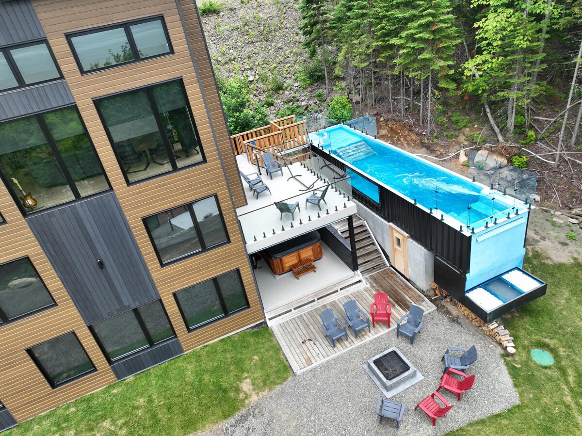 An elevated view of the contemporary chalet reveals a sleek, modern design with expansive windows. A spacious outdoor area features a wooden deck, fire pit, and seating arrangements. The unique container pool is positioned adjacent to the structure, surrounded by natural landscaping.