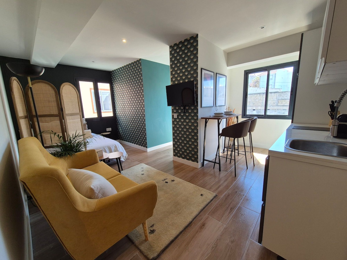 A modern studio apartment features a vibrant yellow sofa alongside a dining area with two high stools. A television is mounted on the wall, and a well-equipped kitchenette is visible. Large windows allow natural light to brighten the space, contrasted by decorative patterned walls.