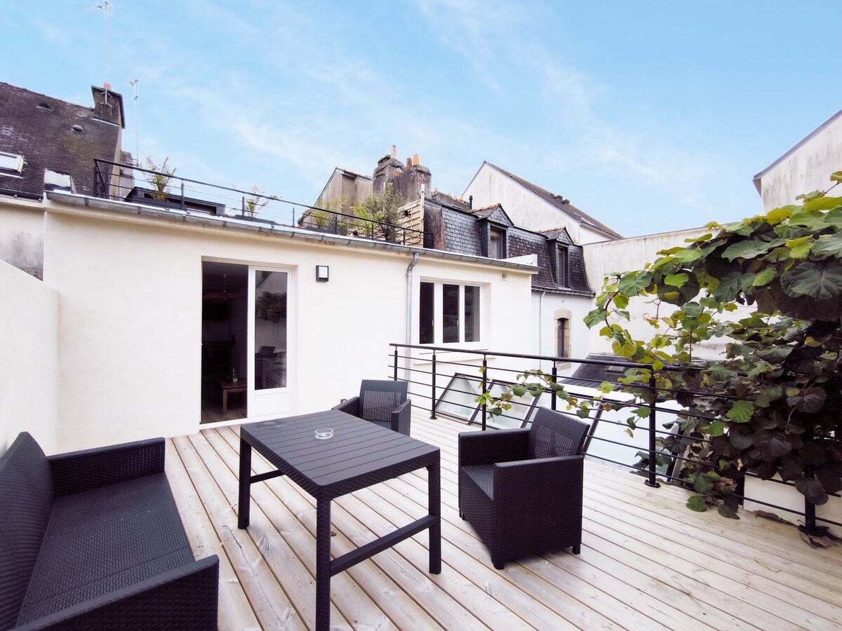 An outdoor terrace featuring a wooden deck and modern black furniture, including two armchairs and a table. Lush greenery is present, providing a natural touch. Access from the interior is visible through a sliding glass door.