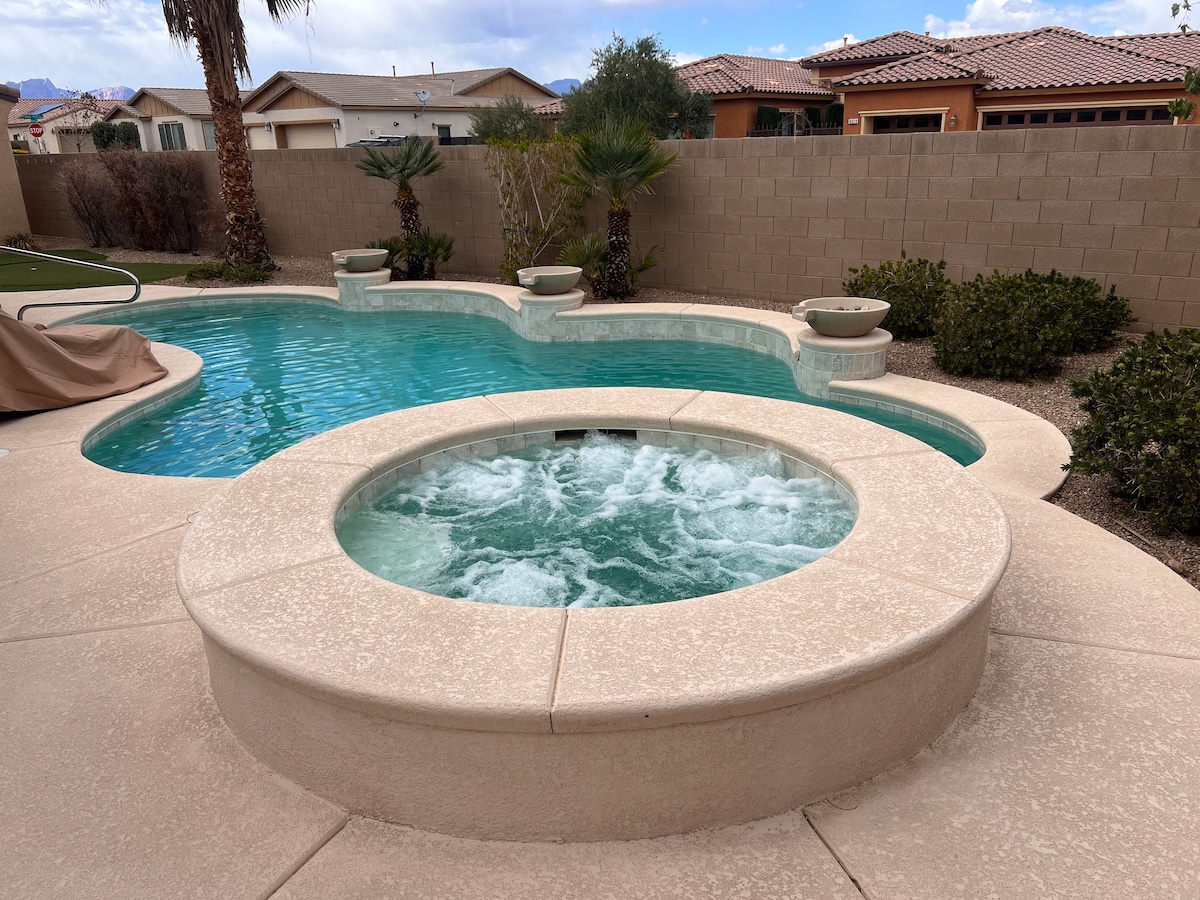 A serene pool area is showcased, featuring a spacious swimming pool with a relaxing hot tub positioned nearby. Surrounding landscaping includes palm trees and well-maintained shrubs, providing a sense of privacy. The neutral-toned patio enhances the tranquil ambiance of the outdoor space.
