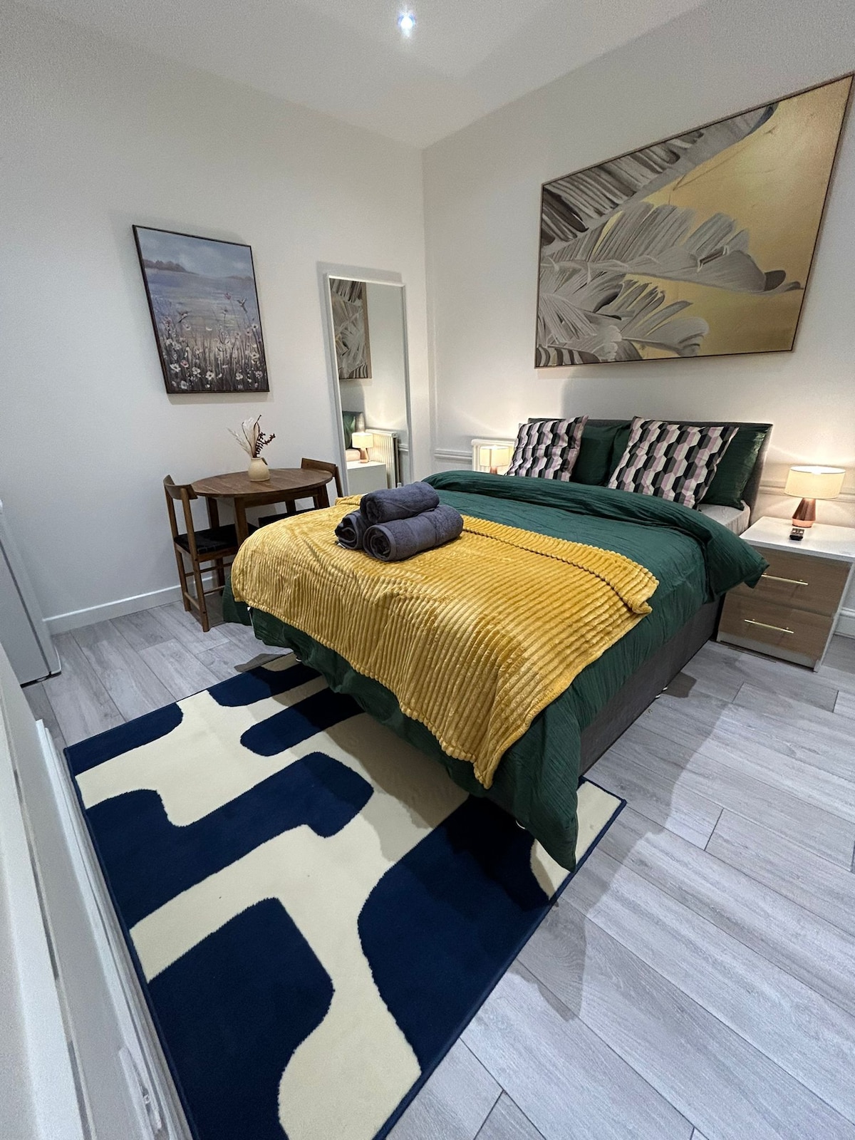 A comfortable room is furnished with a spacious bed layered with a textured yellow throw and neatly arranged towels. A small wooden table is positioned near the bed. Wall art and a mirror add to the decor, while a blue and cream patterned rug covers the floor.